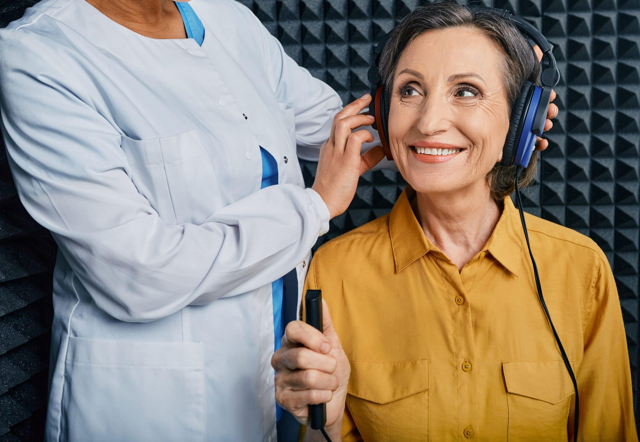 Portrait senior woman with white toothy smile while hearing check-up with ENT-doctor at soundproof audiometric booth using audiometry headphones and audiometer