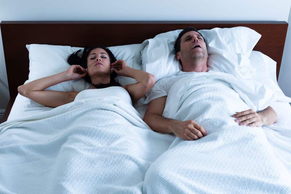 A heterosexual couple in bed. The man on the right has his mouth open as though snoring. The woman on the left is placing ear plugs into her ears.