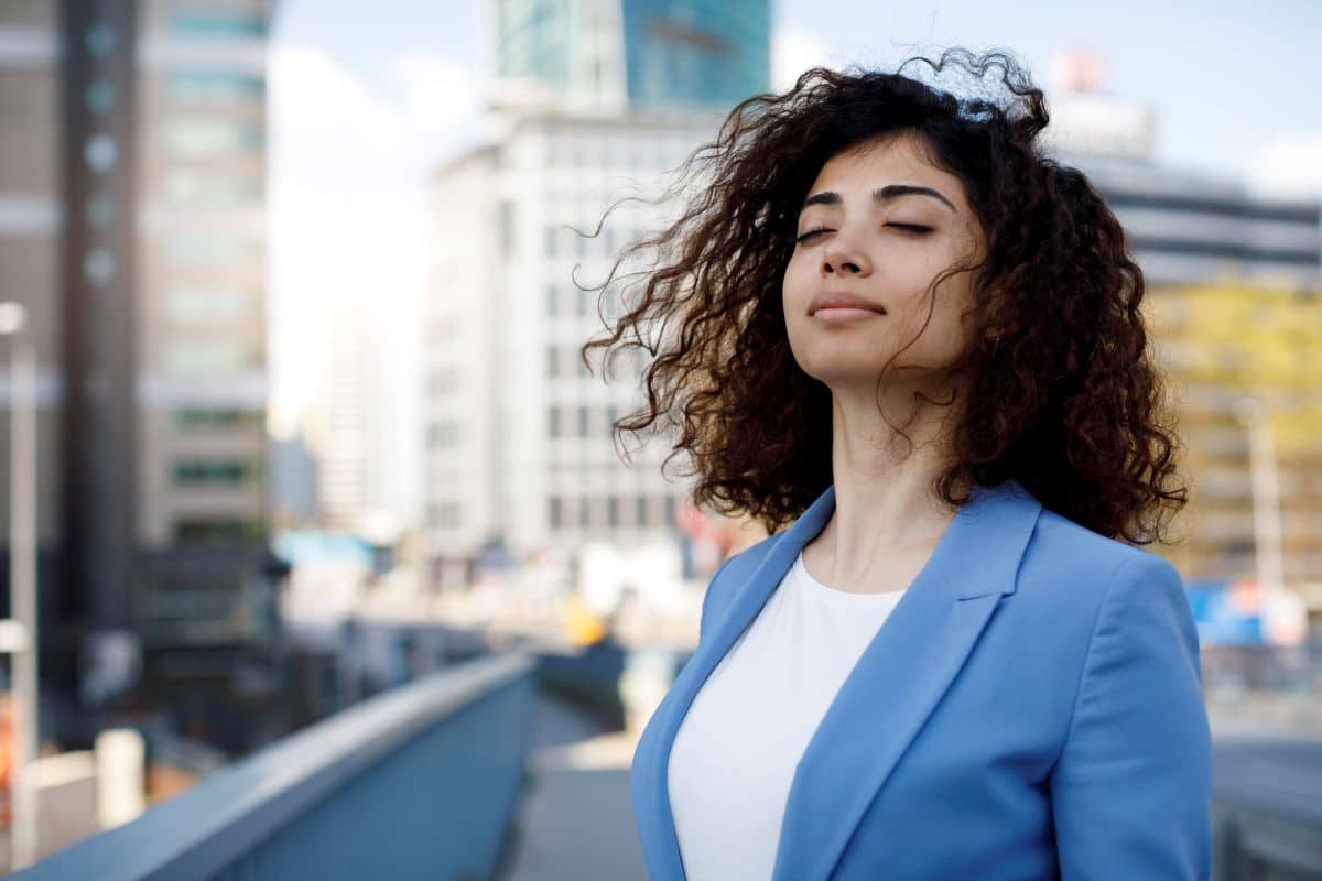 younger woman in an outdoor cityscape closing her eyes as the wind blows her hair forward